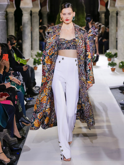 The model walks the runway in an open-front long-sleeve coat with a multi-colored floral design, layered over a cropped bustier top with the same floral design and white trousers. Behind her, another model walks in the opposite direction, with the blurred background revealing the audience and potted plants at the base of tall arches.