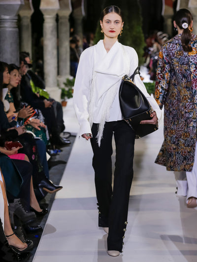 Holding a black duffle bag, the model walks the runway in a white long sleeve blouse with fringe detailing and black trousers. Behind her, another model walks in the opposite direction, with the blurred background revealing the audience and potted plants at the base of tall arches.