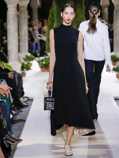 Holding a black cube-like handbag with a cutout floral design, the model walks the runway in a black sleeveless mock-neck knit dress. Behind her, another model walks in the opposite direction, with the blurred background revealing the audience and potted plants at the base of tall arches.