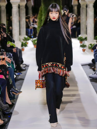 Holding an orange cube-like handbag with a floral design, the model walks the runway in a black long-sleeve turtleneck knit pullover with floral embroidery and fringe trim, paired with black trousers. Behind her, another model walks in the opposite direction, with the blurred background revealing the audience and potted plants at the base of tall arches.