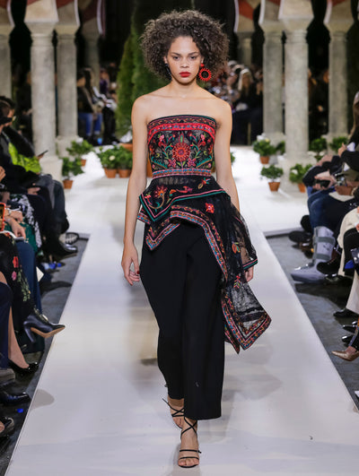 The model walks the runway in a black strapless semi-sheer blouse with multi-colored floral embroidery and an asymmetrical hemline. Behind her, the blurred background reveals the audience and potted plants at the base of tall arches.