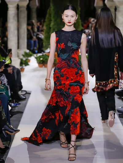 The model walks the runway in a black sleeveless trumpet evening gown with multi-colored textured floral embroidery. Behind her, another model walks in the opposite direction, with the blurred background revealing the audience and potted plants at the base of tall arches.