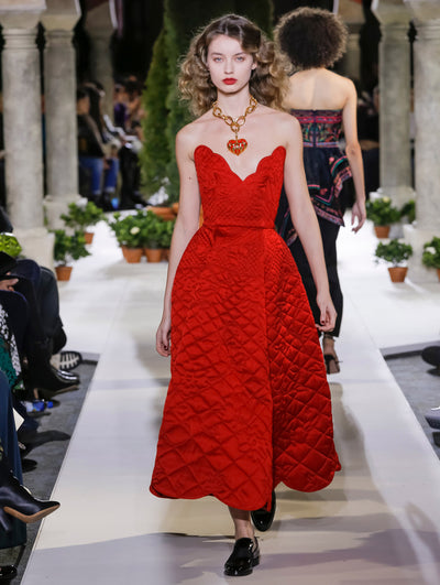 The model walks the runway in a red strapless V-neck cocktail dress with a quilted design. Behind her, another model walks in the opposite direction, with the blurred background revealing the audience and potted plants at the base of tall arches.