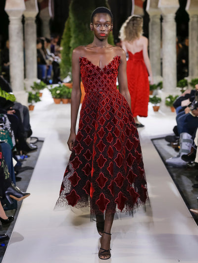 The model walks the runway in a strapless V-neck burgundy velvet cocktail dress with a vintage-inspired design. Behind her, another model walks in the opposite direction, with the blurred background revealing the audience and potted plants at the base of tall arches.