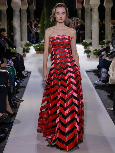The model walks the runway in a black and red striped strapless evening gown. Behind her, the blurred background reveals the audience and potted plants at the base of tall arches.