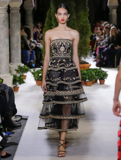 The model walks the runway in a black strapless cocktail dress with a gold threadwork embroidered design and tiered skirt. Behind her, the blurred background reveals the audience and potted plants at the base of tall arches.