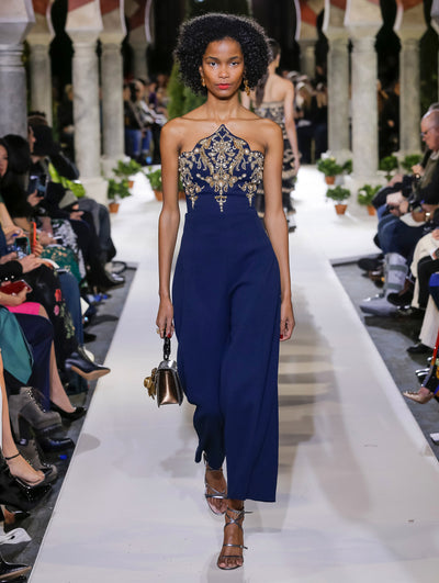 Holding a metallic gardenia appliqué handbag, the model walks the runway in a blue strapless jumpsuit with a gold threadwork embroidered design concentrated on the bodice. Behind her, another model walks in the opposite direction, with the blurred background revealing the audience and potted plants at the base of tall arches.