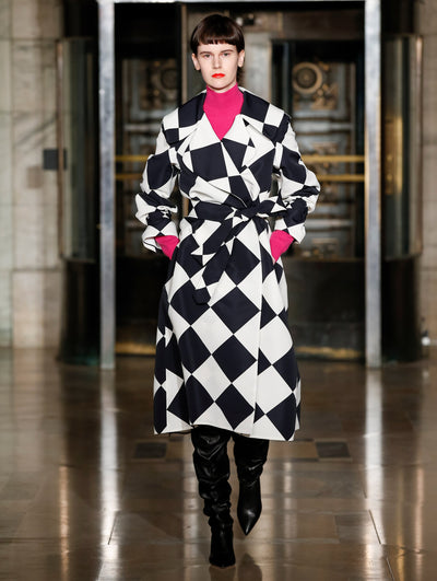 A model wears a black and white coat with geometric shapes and pink turtleneck