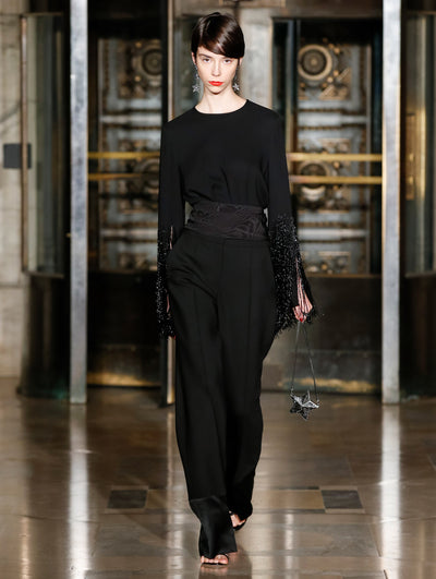 A model wears black pants and a black top with sparkly black fringe at the sleeve