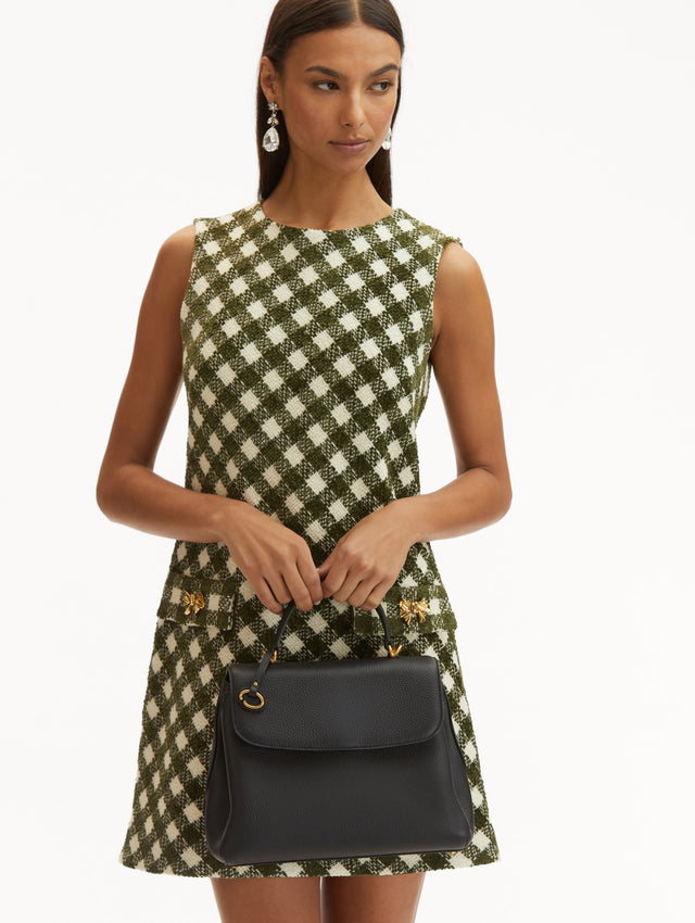 Model in plaid dress holding The Poppy leather bag in dark navy with O logo tag around the top handle. Front View