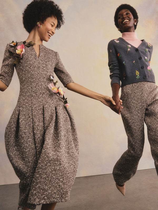 The two models are in motion, smiling, and holding hands. The model on the left is wearing a tweed dress with floral accents. The model on the right is wearing a pink turtleneck, a floral embroidered cardigan, and tweed trousers.