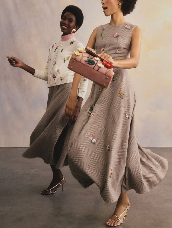 The two models are walking in motion side-by-side. The model on the right is wearing a pink turtle-neck sweater, a white cardigan with floral embroidery, and a midi skirt. The model on the right is wearing a dress with floral accents and is holding a matching mini bag.