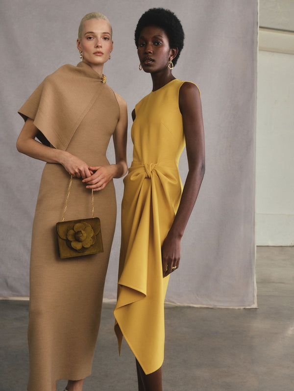 The two models are posed standing side-by-side. The model on the left is wearing a tan dress with cape detail and is holding an evening bag with a flower accent. The model on the right is wearing a yellow dress.