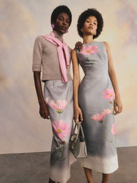 The two models are posed side-by-side, leaning towards each other. The model on the right is wearing a camel turtle-neck sweater, a grey skirt with a pink flower design, and a pink cardigan tied around the neck. The model on the right is wearing a grey dress with a pink flower design.