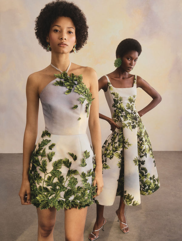 The two models are posed with one in front of the other. The model on the left is wearing a mini, halter dress with a sky, cloud print and leaf detailing. The model on the right is wearing a midi dress with a sky, cloud print and leaf detailing.