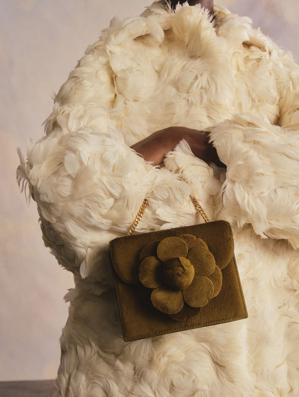 The model is wearing an ivory feather coat and is holding an olive-green mini bag with a flower detail.