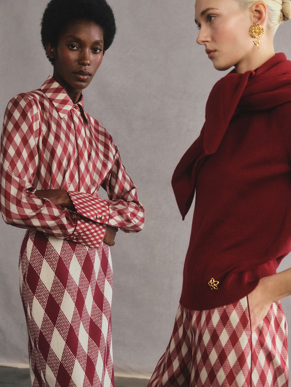 The two models are posed facing towards each other. The model on the left is wearing a silk, collared button down in a red and white argyle print, with a matching silk argyle skirt. The model on the right is wearing a red sweater with a matching scarf, and silk, red and white argyle trousers.