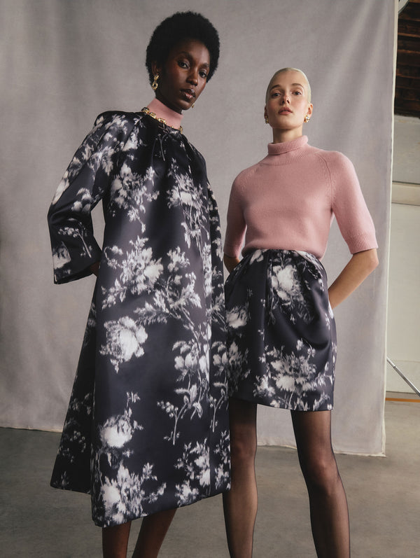 The two models are posed standing beside each other. The model on the left is wearing a long sleeve peony print satin coat. The model on the right is wearing a peony print satin skirt and a pink quarter-sleeve turtle-neck top. 
