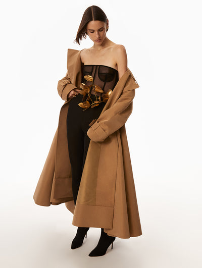 Full body view of model wearing black jumpsuit with gold leaf accent around waist. Model is also wearing brown jacket over the jumpsuit. 