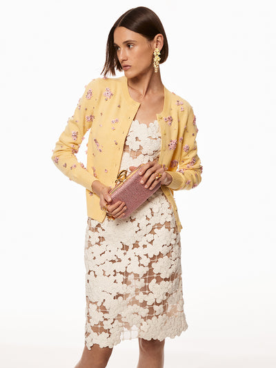 Model wearing white floral grid dress. Model is wearing a yellow sweater with pink pink floral embroidery and holding a pink crystal clutch with braided "O" emblem on top. 