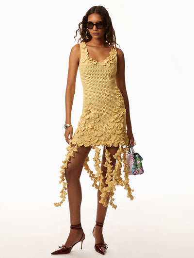 Model wearing a yellow dress with floral accents around neckline and cascading from hemline. Model is holding a pink floral printed hang bag with a blue back round. Model is wearing sunglasses with gold hoop earrings and a gold bracelet. 