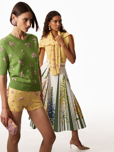First model is wearing a green three quarter length sweater with pink floral embroidery and yellow shorts with pink floral embroidery, she is holding a pink floral clutch and wearing pink floral pom pom earrings. The second model is wearing a white and green skirt with printed florals and a yellow tank with a yellow sweater with pink floral embroidery draped over shoulders and has white and gold triple drop earrings.  