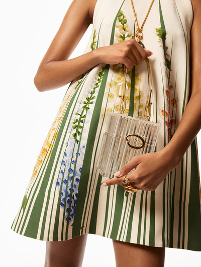 Model is wearing white dress with green slits and printed foxgloves, she is holding plexiglass bag with gold Oscar "O" logo and gold strap. Model is wearing gold rings and a gold necklace. 