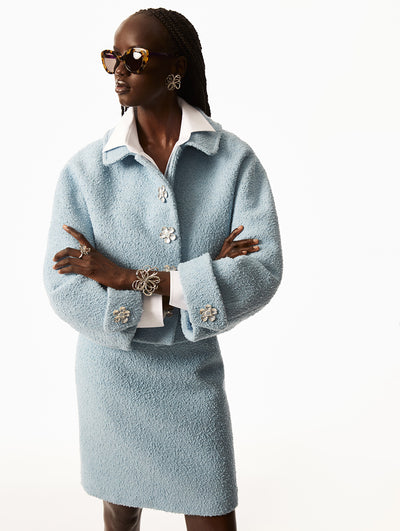 Model is wearing blue skirt with white long sleeve underneath a blue jacket silver floral button detailing. Model is wearing silver wired flower bracelet, ring and earrings. 