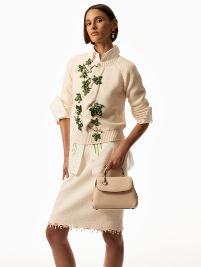 Model is wearing ivory long sleeve sweater with green leaf embroidery with a white tweed skirt with pearl drops along the hemline. Model has white button down underneath the sweater and is holding a beige mini poppy hand bag. 