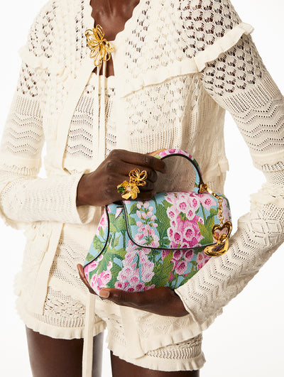 Model is wearing a white lace sweater and shorts with gold wire flower jewelry accents while holding a hang bag with pink printed foxgloves on a blue back round and a gold bag charm. 