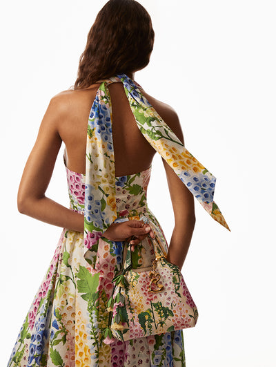 Back view of model wearing pink, yellow, blue and green foxglove dress with self-tie neck. Model is holding a matching handbag with a gold bag charm. 