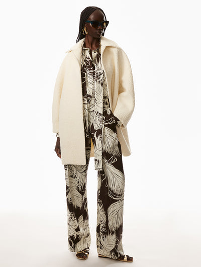 Model is wearing a brown jumpsuit with printed white feather detailing and a white jacket. Model is wearing gold wire earrings. 
