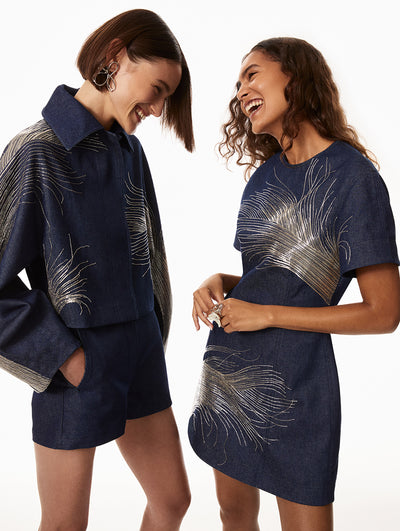 The first model is wearing a blue denim jacket with silver embroidery detailing and matching denim shorts with silver floral earrings. The second model is wearing blue denim dress with silver embroidery detailing and a silver floral ring. 