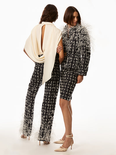Back view of the first model wearing ivory top draped over shoulders and dark indigo woven denim pants with white feathers. The second model is wearing a dark indigo denim woven jacket with a gold emblem on belt and skirt with white feather on the bodice. 