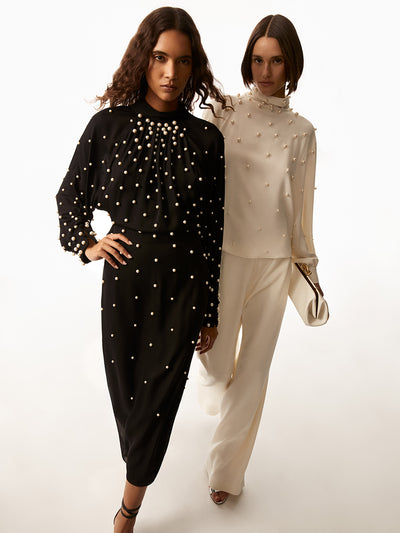 First model in the photo is wearing a long sleeve black dress with pearls scattered across. The second model is wearing a ivory top with pearls and ivory pants and holding a white clutch hand bag.  