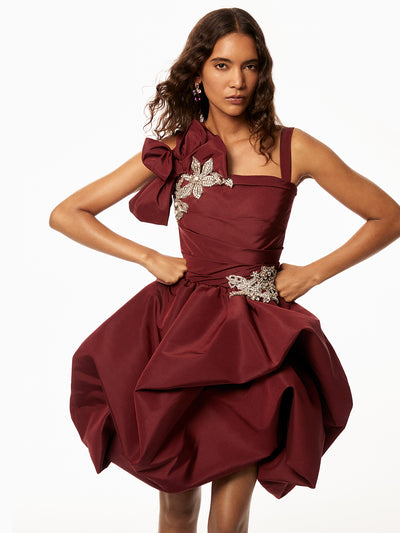Model is wearing merlot dress with silver floral detailing on shoulder and waist with a bow accent on the shoulder. 