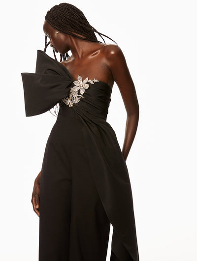 Model wearing black top with bow detail on shoulder and silver floral embroidery and black pants. 