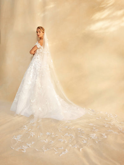 Model is wearing and off the shoulder wedding gown with embroidered floral details and long head piece. back view