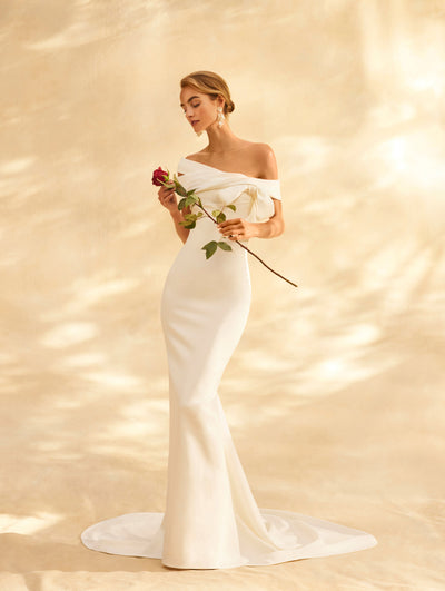 The model is wearing an off the shoulder wedding dress with a bow detailing. 