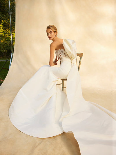 The model is wearing a strapless wedding dress with a large bow on the back while sitting in a chair. 