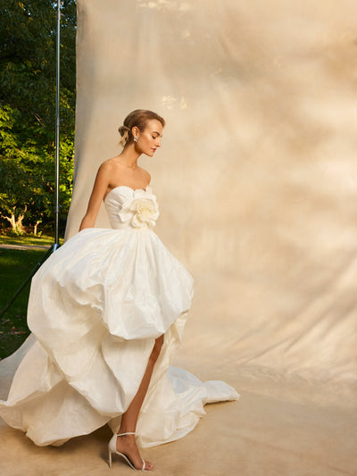 The model wears a high low wedding gown with a bubble skirt and three-dimensional flower in the center.