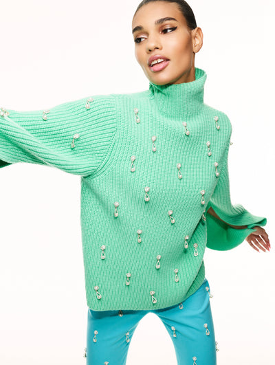A model poses against a white backdrop in an Oscar de la Renta green sweater and blue trousers with embroidered detail. 
