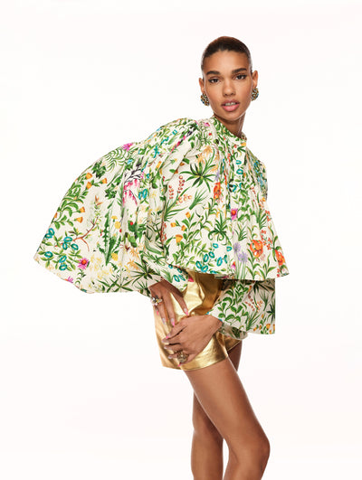 A model poses against a white backdrop in an Oscar de la Renta floral blouse with gold shorts. 