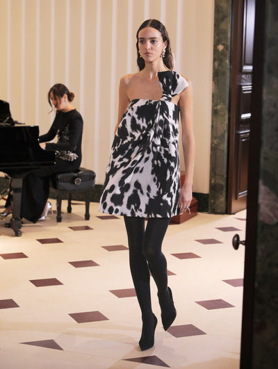 The model wears a one-shoulder cow print cocktail dress paired with black tights and a black clutch.  