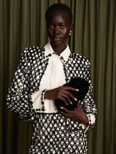 The model wears a black and white embroidered jacket and mid-length skirt set. The look is paired with a white collared blouse.  