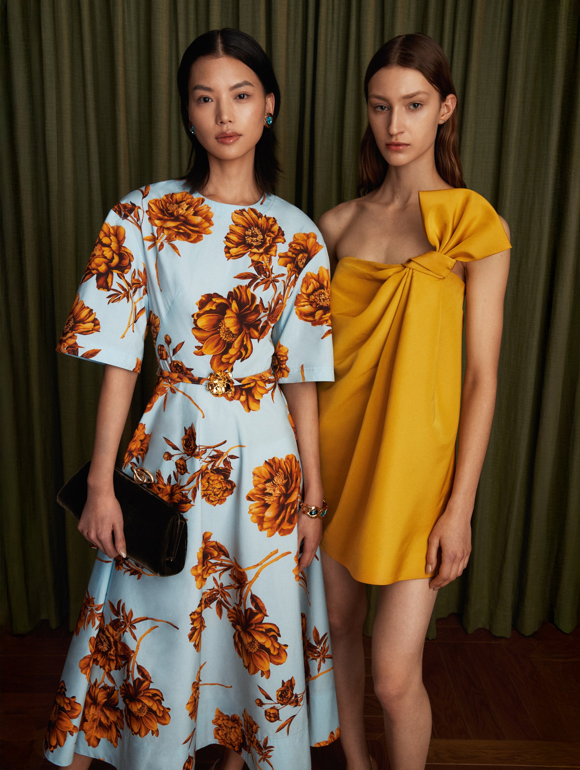 The model wears a cotton poplin short sleeve midi dress. The print of the dress features the flower of the season: peonies. The flower is mustard, and the background is light blue.  The model on the right is wearing a yellow one shoulder mini dress with a bow detailing. 