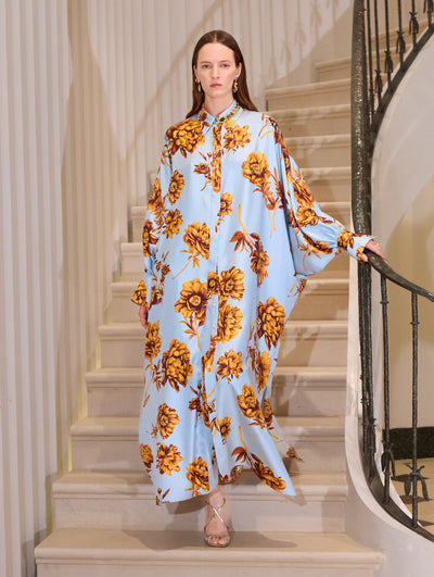 The model wears a peony printed caftan in mustard and light blue open with light blue pants and sandals.  