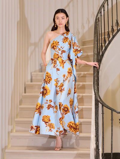 The model wears a cotton poplin one-shoulder midi dress paired with a belt. The print of the dress features the flower of the season: peonies. The flower is mustard, and the background is light blue.  