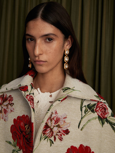 The model is wearing a white jacquard coat with floral motifs and gold hanging earrings. 
