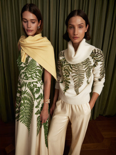 The model on the left is wearing a sleeveless mid-length day dress with a fern motif. The look is styled with a yellow scarf. The model on the right is wearing a white turtleneck with a fern motif. 